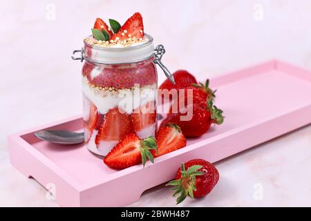 Erdbeerfrucht-Dessert mit Joghurt, gesunden Chia-Samen und gepufften Quinoa-Körnern, die in einem Glas von Beeren umgeben sind, auf einem rosa Holztablett Stockfoto