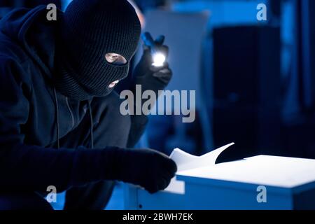 Maskierter Eindringling, der vertrauliche Dokumente hält und liest Stockfoto