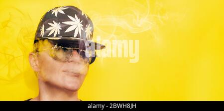 Mädchen in Sonnenbrille und Mütze mit Blättern von Marihuana raucht auf gelbem Hintergrund Stockfoto