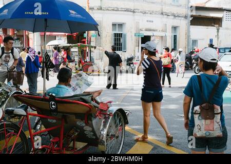 Ein Rikscha-Fahrer liest das Papier im Schatten und wartet auf eine Fahrt mit Touristen, die in Georgetown, Penang, Malaysia vorbeilaufen Stockfoto