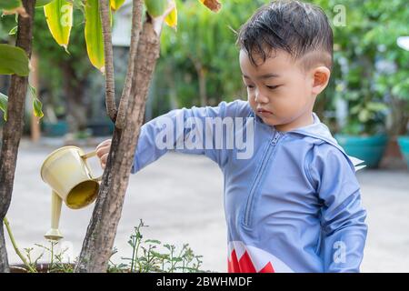 Asiatische niedlich liebenswert kleinen Kleinkind junge in Bewässerung Bäume Topf mit kleinen Dose im Freien. Spaß Baby Gartenarbeit Pflanze im Hinterhof Land Hütte auf Stockfoto