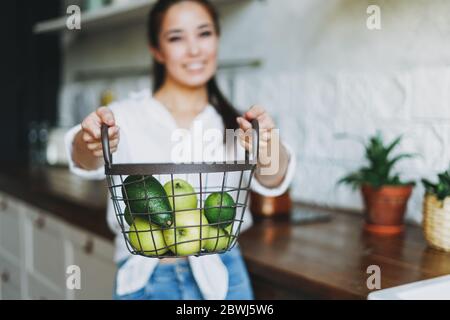 Junge asiatische Frau in weißen Hemd halten Korb mit grünen Früchten und Gemüse in den Händen auf Küche Stockfoto