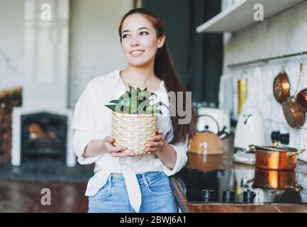 Junge asiatische Frau in weißen Hemd halten Korb mit grünen Haus Pflanze in Weidenkorb in Händen in der Küche Stockfoto