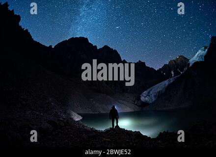 Mann mit seinem Kopflicht schaut in sternenstarker Nacht auf den See die Berge Stockfoto