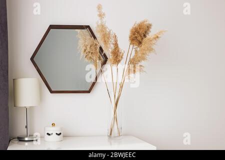 Trockenes Schilf in einer transparenten Glasvase, die neben dem Holzspiegel steht. Stockfoto