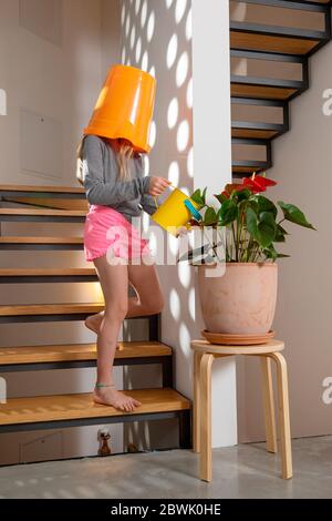 Kleines Mädchen mit Eimer auf dem Kopf kümmert sich um die Blumen ihres Hauses, die Treppe ist aus Holz. Stockfoto