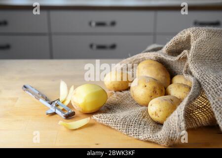 Kartoffeln in einem Sackleinen, eine Hälfte neben einem sehr alten Sparschäler auf einem hölzernen Küchentisch geschält, Kopierraum, ausgewählter Fokus, schmale Tiefe des fie Stockfoto