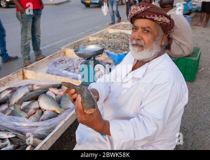 Älterer omanischer Fischhändler, der einen Fisch auf dem Fischmarkt am Straßenrand in Muscat, Sultanat von Oman zeigt. Stockfoto