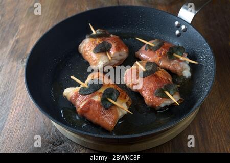 Gebratenes Hähnchen Saltimbocca mit luftgetrockneten Schinken und Salbeiblättern in einer schwarzen Pfanne auf einem rustikalen Holztisch, ausgewählter Fokus, schmale Schärfentiefe Stockfoto