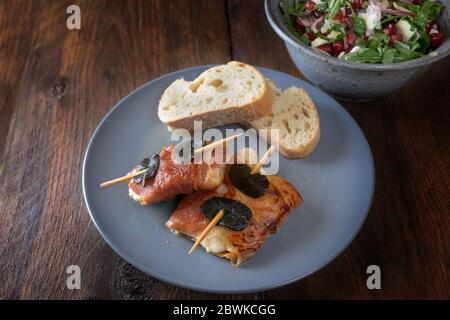 Gebratenes Hähnchen Saltimbocca mit Schinken und Salbeiblättern umwickelt, serviert mit Brot und Salat auf einem dunklen rustikalen Holztisch, ausgewählte Fokus, nar Stockfoto