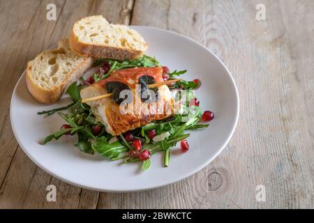 Gegrilltes Hähnchen Saltimbocca mit Schinken und Salbeiblättern umwickelt, serviert mit Brot auf Rucola-Salat auf einem weißen Teller auf einem rustikalen Holztisch, Stockfoto