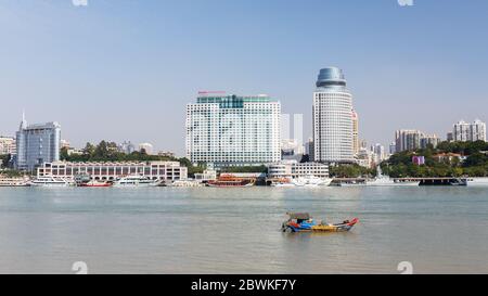 Stadtbild von Xiamen mit kleinem, farbenfrohen Boot, das im Vordergrund vorbeifährt. Symbol für das Zusammenleben von Tradition und Moderne. Stockfoto