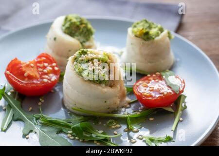 Drei Fischfiletrollen aus Scholle, gefüllt mit Sellerie und Rucola, serviert mit gegrillten Tomaten, Rucola-Salat und Sesam auf einem blauen Teller, Stockfoto