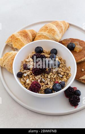 Gesunde und nahrhafte Frühstücksplatte mit Müsli in einer weißen Schüssel mit Haferflocken Pfannkuchen und Croissants serviert an seiner Seite in einer runden weißen Keramik Stockfoto