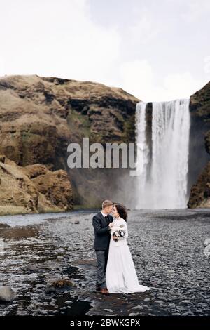 Hochzeit in Island. Hochzeitspaar in der Nähe des Skogafoss Wasserfalls. Die Braut und der Bräutigam umarmen sich am Fluss. Stockfoto