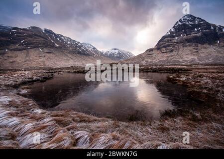 Ein kleiner gefrorener See steht in frostbedeckter Moorlandschaft und unter schneebedeckten Bergen am Rannoch Moor im West Highlands von Schottland. Stockfoto