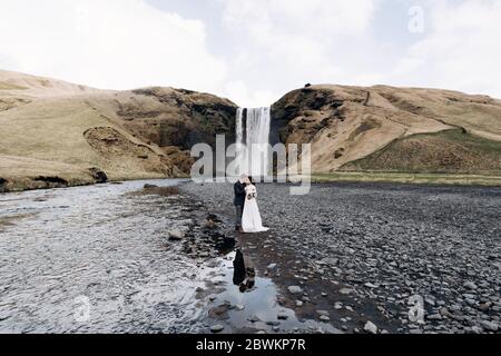 Hochzeit in Island. Hochzeitspaar in der Nähe des Skogafoss Wasserfalls. Die Braut und der Bräutigam umarmen sich am Fluss. Stockfoto