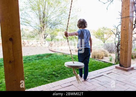 6 Jahre alter Junge mit einer Seilschaukel auf einer großen Veranda im Schatten. Stockfoto