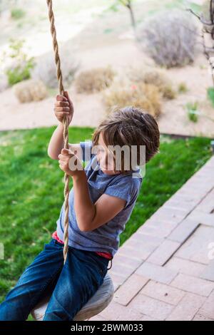 6 Jahre alter Junge mit einer Seilschaukel auf einer großen Veranda im Schatten. Stockfoto