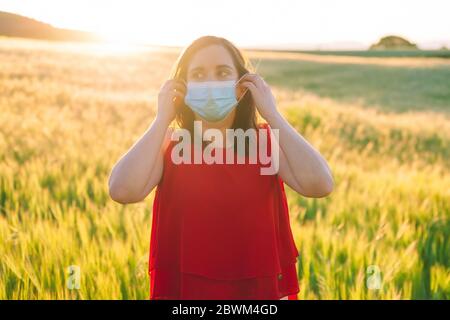 Glücklich fröhlich junge Frau Entfernen medizinischen Gesichtsmaske, auf der Wiese im Freien während Sonnenuntergang im Frühling stehen. Konzept des Endes der Coronavirus-Pandemie . Hochwertige Fotos Stockfoto