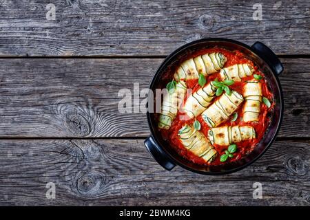 Italienische Zucchini Rollatini gefüllt mit weichem Ricotta, Baby Spinat und Basilikum Blätter in Tomate Passata gebacken serviert auf einer schwarzen Backform auf einem hölzernen b Stockfoto