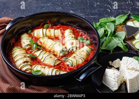 Zucchini-Streifen mit einer Füllung aus Ricotta mit Baby-Spinat gemischt gerollt, frische Basilikumbrüche in Tomatensauce gebacken serviert auf einer runden schwarzen Backform Stockfoto