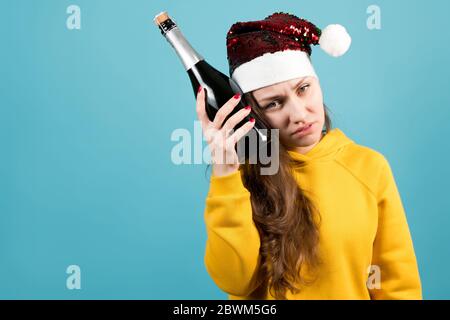 Mädchen in einem gelben Sweatshirt auf blauem Hintergrund mit einem Kater drückt eine kalte Flasche Champagner auf ihren Kopf Stockfoto