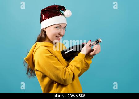 Mädchen in Weihnachtsmütze auf blauem Hintergrund mit einem schlauen Lächeln freut sich auf eine Flasche Champagner zu öffnen Stockfoto