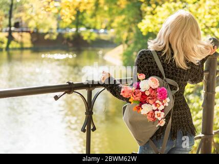 Foto von der Rückseite der jungen blonden verträumten Frau mit Blumenstrauß in grauem Rucksack in einem Frühlings-Sommerpark Stockfoto