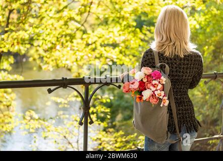 Foto von der Rückseite der jungen blonden verträumten Frau mit Blumenstrauß in grauem Rucksack in einem Frühlings-Sommerpark Stockfoto