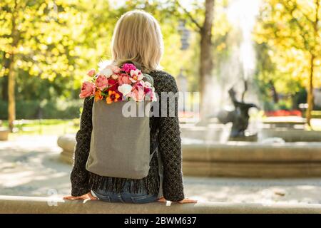 Foto von der Rückseite der jungen blonden verträumten Frau mit Blumenstrauß in grauem Rucksack in einem Frühlings-Sommerpark Stockfoto