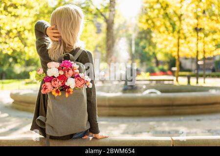 Foto von der Rückseite der jungen blonden verträumten Frau mit Blumenstrauß in grauem Rucksack in einem Frühlings-Sommerpark Stockfoto