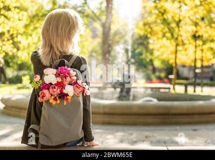 Foto von der Rückseite der jungen blonden verträumten Frau mit Blumenstrauß in grauem Rucksack in einem Frühlings-Sommerpark Stockfoto