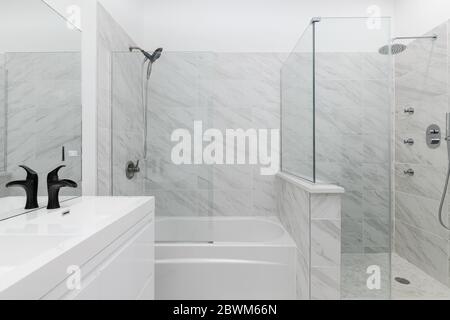 Ein ganz weißes Hauptbadezimmer mit schwarzen Armaturen und Armaturen, die den Waschtisch und die Dusche bei geöffneter Tür zeigen. Stockfoto