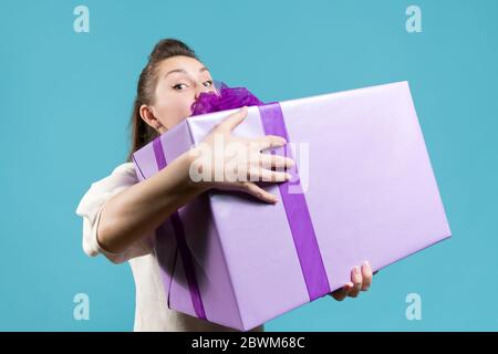 Ein Mädchen späht hinter einem riesigen Geschenk, das sich festklammert. Blauer Hintergrund, Studio Stockfoto