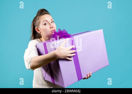 Mädchen hält ein großes Geschenk, es an sich selbst zu halten. Das Mädchen ist ein wenig traurig, direkt auf die Kamera schauend. Blauer Hintergrund, Studioaufnahme Stockfoto