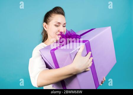 Das Mädchen hält ein großes Geschenk, hält es an sich fest und lächelt warm und schaut ihn an. Blauer Hintergrund, Studioaufnahme Stockfoto