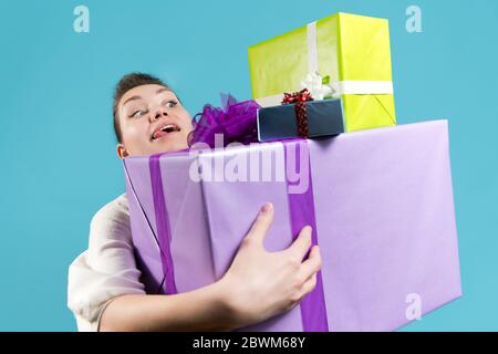 Die junge Frau blickt voller Freude und Vorfreude auf die Geschenke, die sie in ihren Händen hält. Blauer Hintergrund, Studio Stockfoto