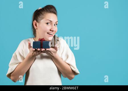 Junge Frau hält ein kleines Geschenk auf ihren Handflächen nahe bei sich und lächelt hell. Blauer Hintergrund, Kopierbereich Stockfoto