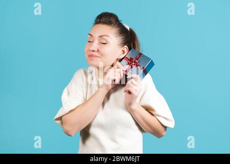 Junge Frau freut sich über ein Geschenk, bringt es ihr ins Gesicht und lächelt. Blauer Hintergrund Stockfoto