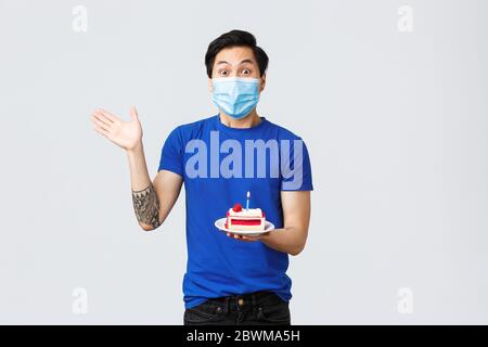 Selbstquarantäne, Home Lifestyle und Feier Konzept. Aufgeregt jungen Mann Geburtstag feiern, asiatische Kerl in medizinischen Maske winken Hand und halten b Stockfoto