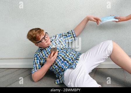 Der rothaarige Junge mit Brille und Smartphone, die auf den Boden gefallen ist, erhält die von einem Erwachsenen angebotene OP-Maske. Junge mit karierten Shirt berührt die s Stockfoto