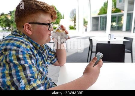 Niedlicher rothaariger Junge mit Handy und Brille trinkt im urbanen Kontext Eiswasser mit Zitrone. Der Junge mit der Brille schaut auf den Bildschirm eines Smars Stockfoto