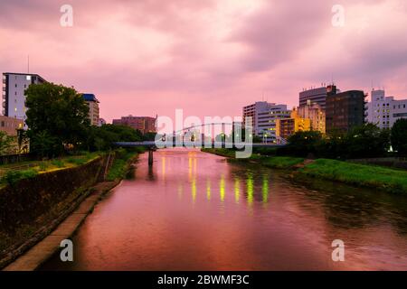 Kumamoto, Japan. Blick auf eine Brücke über den Shirakawa Fluss in Kumamoto, Japan während der Nacht mit Wolkenkratzern Stockfoto