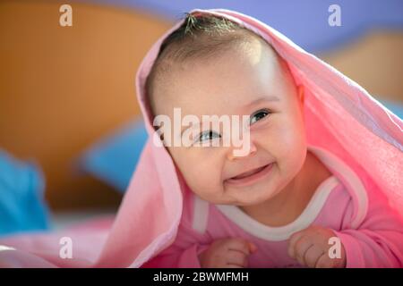 Nette drei Monate alte Baby Mädchen Säugling auf einem Bett auf ihrem Bauch mit Kopf nach oben in die Kamera mit ihren großen Augen suchen. Natürliches Licht im Schlafzimmer. Stockfoto