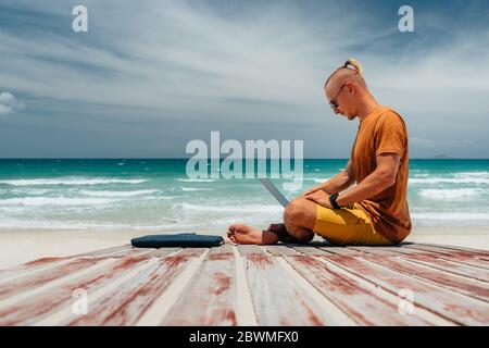 Junger Mann mit langen Haaren am Meer an einem sonnigen Tag, arbeitet mit seinem Laptop, arbeitet aus der Ferne mit einem Unternehmen. Bloner, Freiberufler, arbeitet durch das I Stockfoto