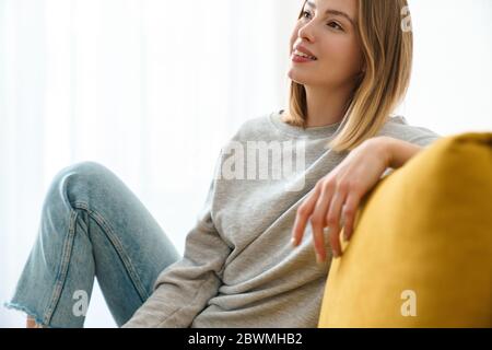 Schöne lächelnde junge blonde Frau in lässigen Kleidern, die zu Hause auf einer Couch ruht und wegschaut Stockfoto