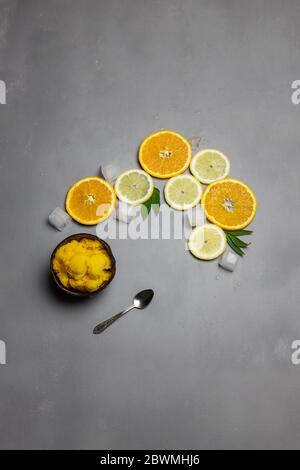 Kokosnussschale voll mit Zitrusfrüchten Sorbet Eis und kleinen metallischen Löffel, in Scheiben geschnittene Orangen und Zitronen auf der Seite mit Eiswürfeln auf grauem Beton Bac Stockfoto