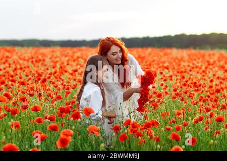 Kleine glückliche Mädchen mit Rotschopf Mutter in weißen Kleidern macht Kranz auf Mohn Feld bei warmen Sommer Sonnenuntergang Stockfoto