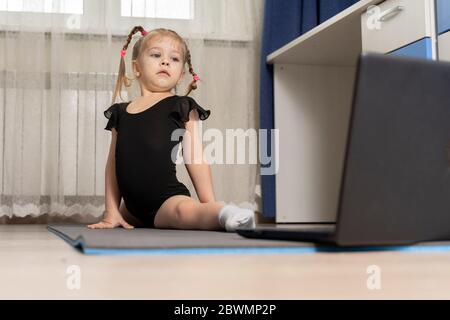 Ein kleines Mädchen in einem schwarzen Turntrikot macht Gymnastik zu Hause online vor einem Laptop. Platz für Text und Kopierer. Stockfoto